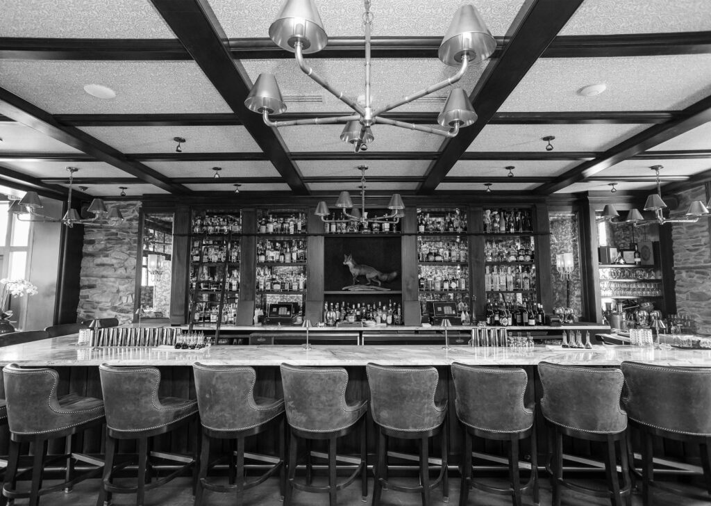 An empty bar with stools, shelves of bottles, and hanging lights, in monochrome.