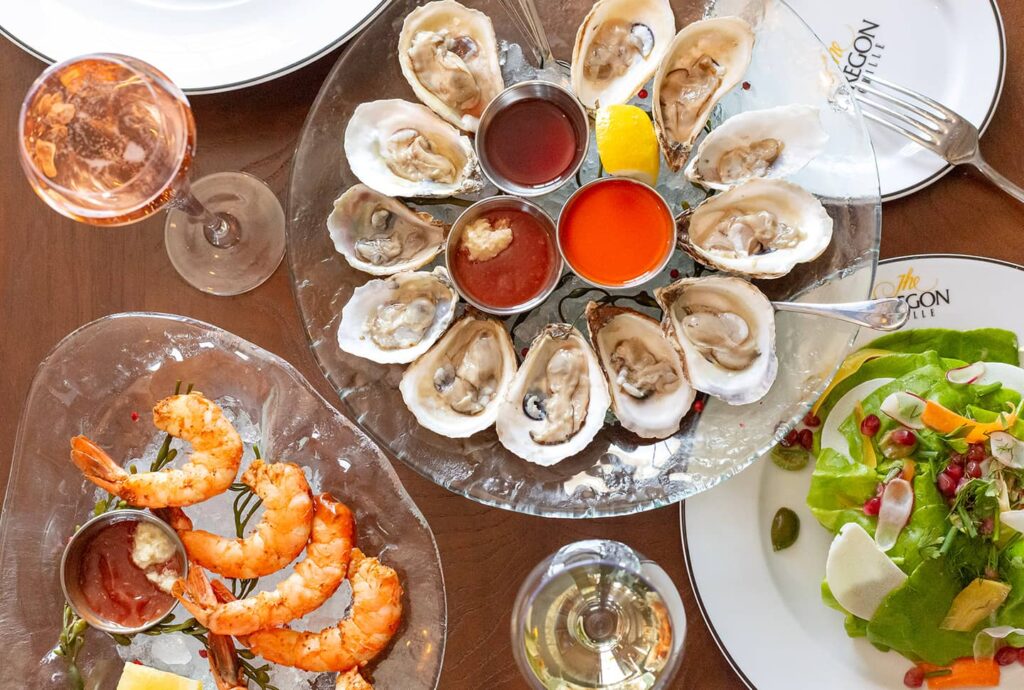 An overhead view of a dining table with a platter of oysters, shrimp cocktail, salad, and glasses of wine.