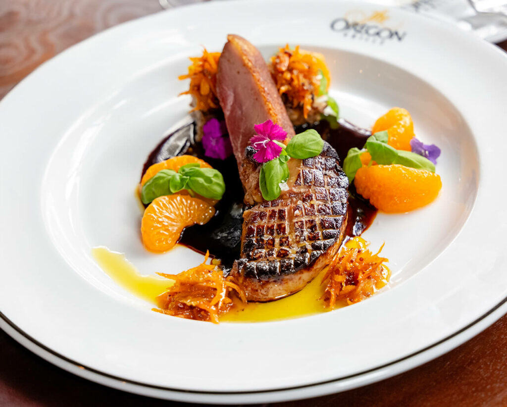 Roasted duck breast served with orange segments and a garnish of edible flowers on an elegant white plate.