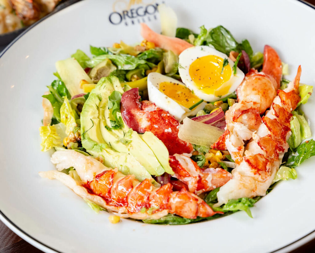 A fresh lobster salad with avocado, mixed greens, and a boiled egg served on a white plate.