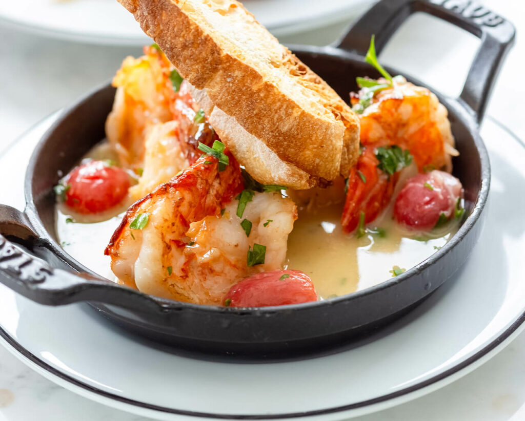 Sautéed shrimp with herbs and tomatoes served in a skillet accompanied by a slice of toasted bread.