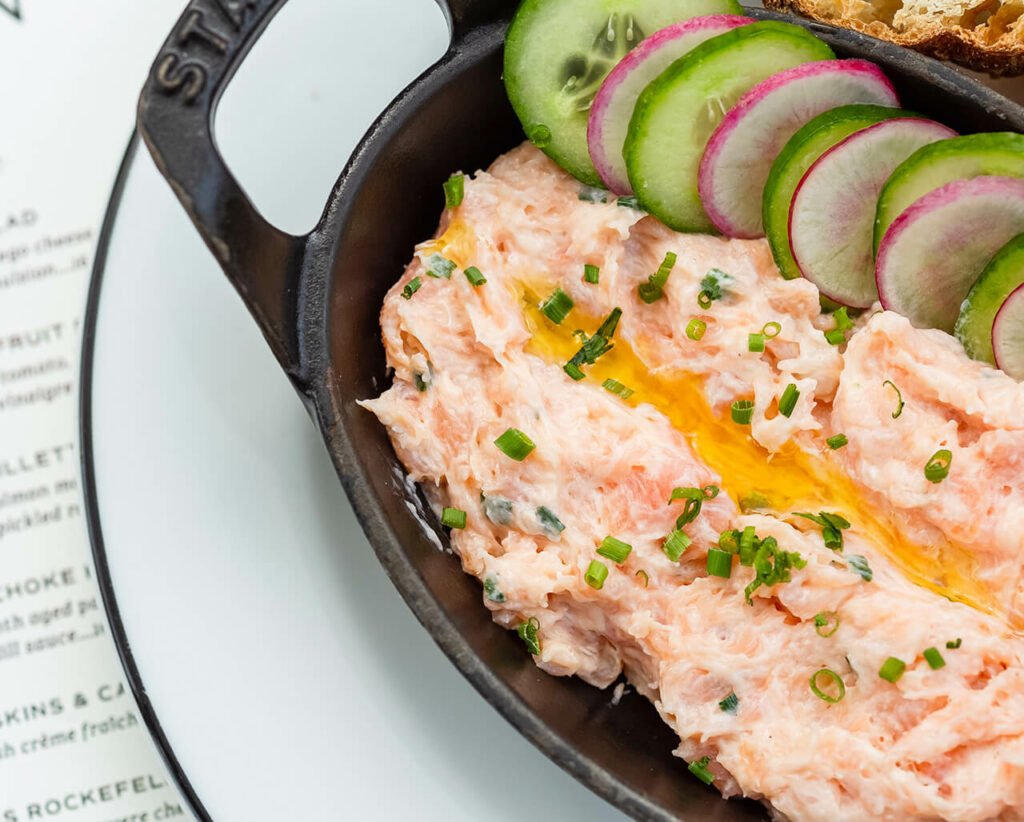A skillet containing salmon dip garnished with chives, served with slices of cucumber and toasted bread, placed on a menu.