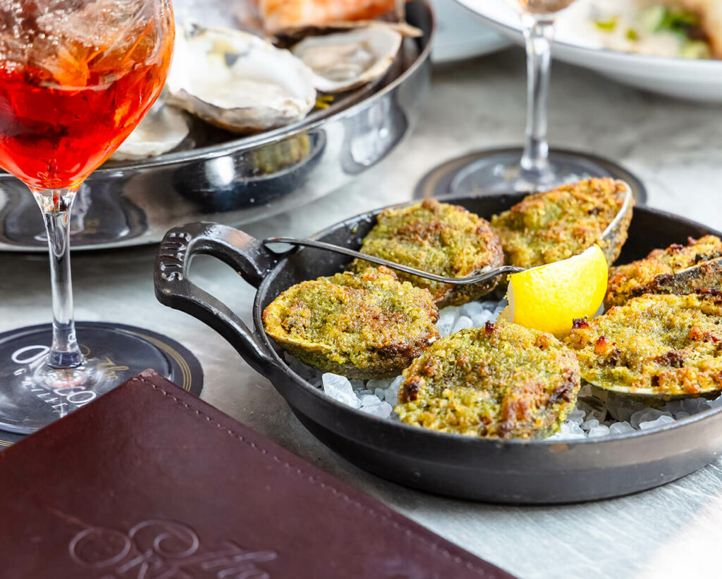 A pan of baked oysters set on a bed of ice with a lemon wedge, accompanied by a glass of wine and more dishes in the background.